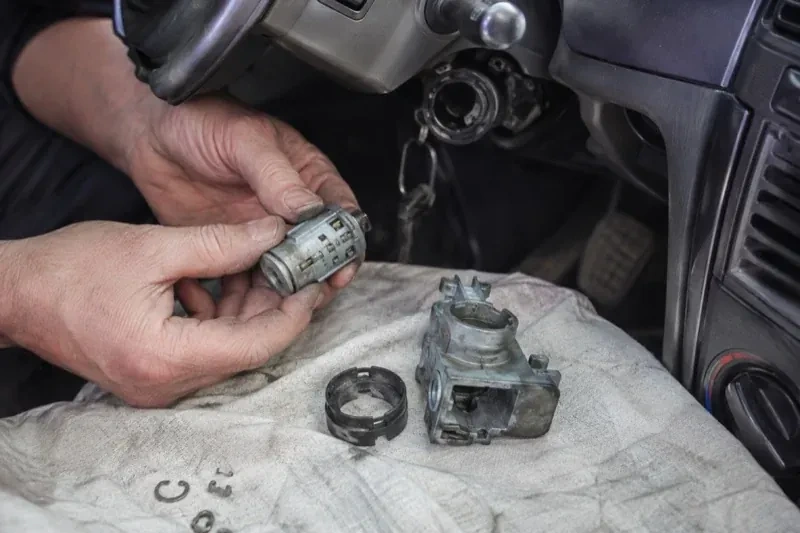 Hands holding a removed ignition cylinder beside a car steering column, with the housing and small parts on a cloth. — Ignition Lock Repair, Springwater